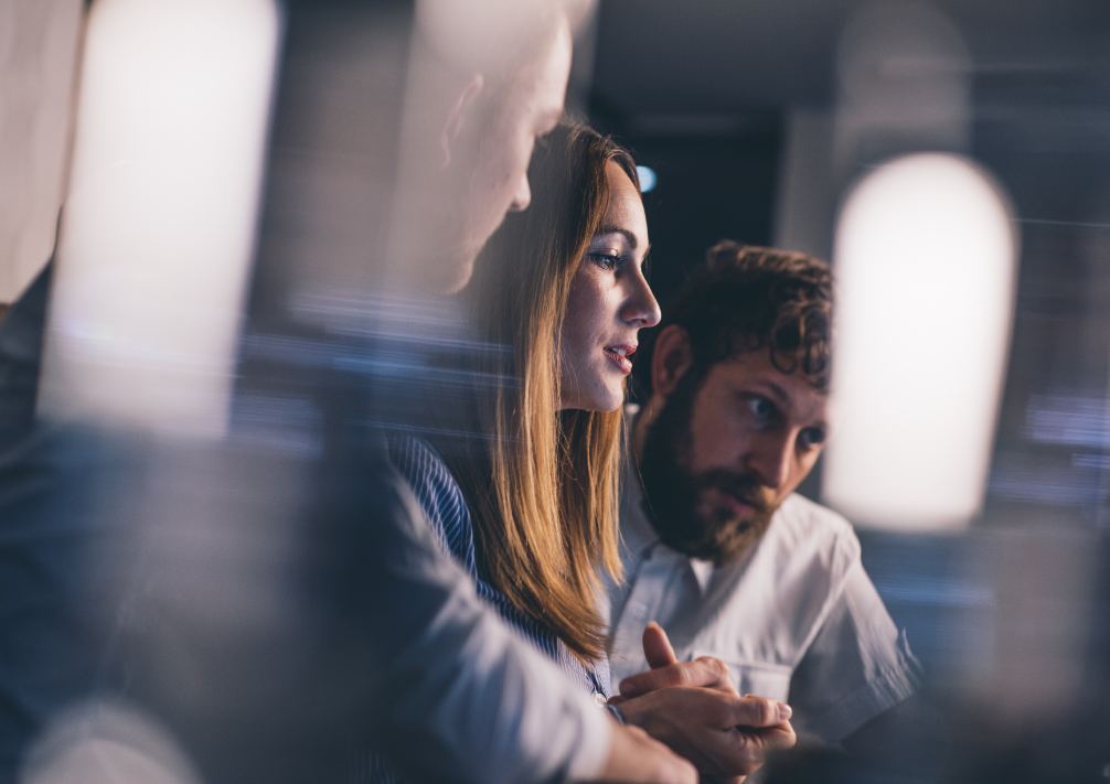 Three people are pictured in a dimly lit setting, engaged in a serious conversation.
